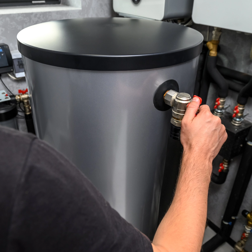 Person adjusting a grey hot water cylinder with connected heating components.